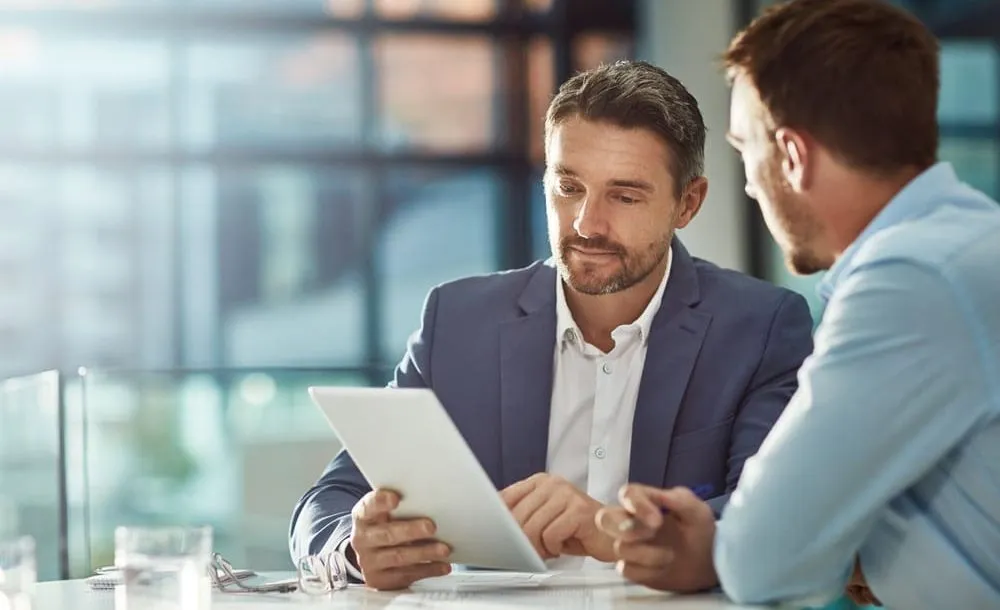Two businessmen, dressed in professional attire, are seated together at a desk in an office setting. One of the businessmen is holding a tablet, displaying an R&D-related document for review, while the other attentively observes. They are discussing and analyzing the contents of the digital document, fostering a collaborative and focused atmosphere indicative of a productive business meeting focused on research and development strategies.