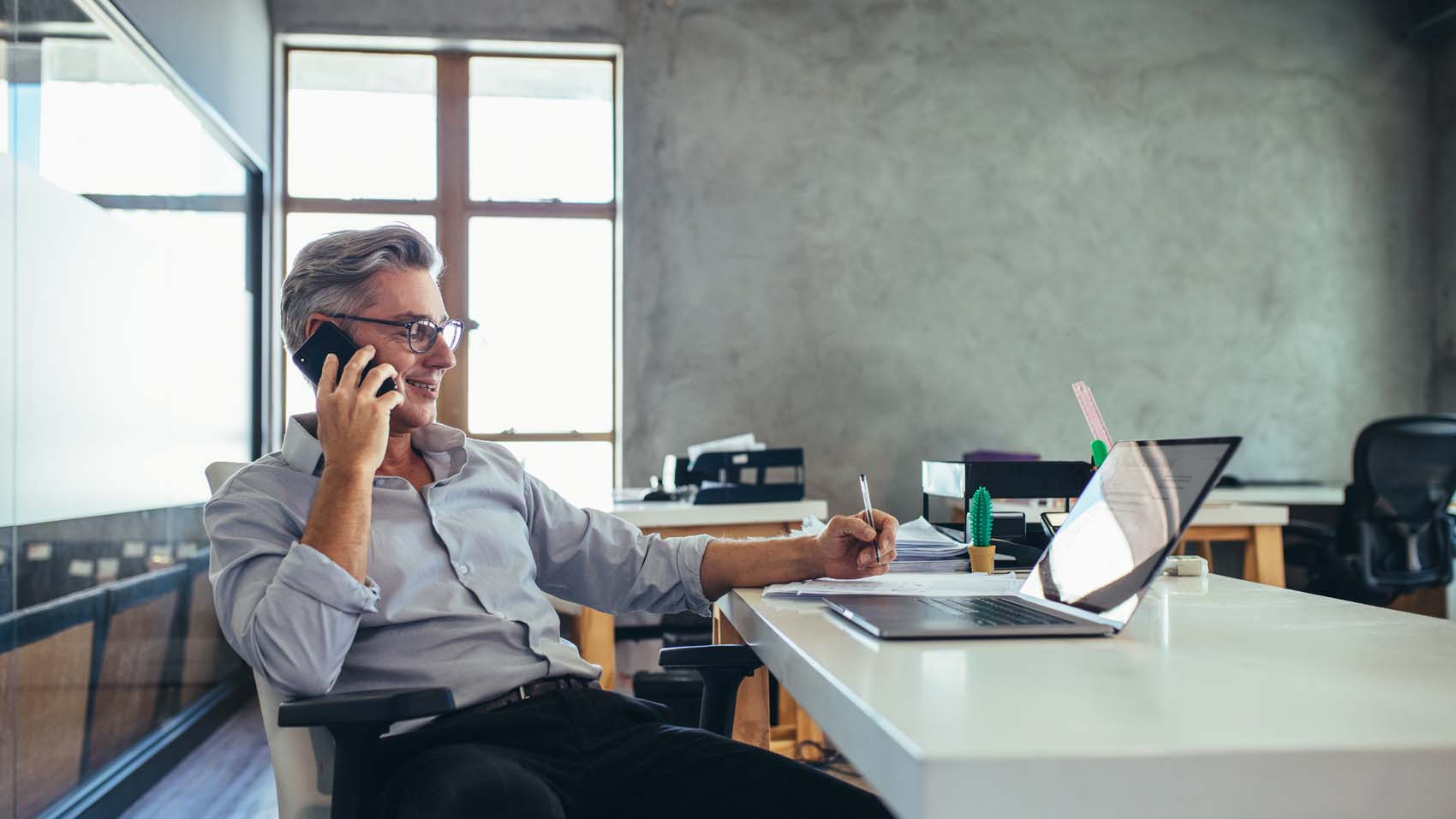 A man sitting at a desk talking on a phone while writing on a sheet of paper.